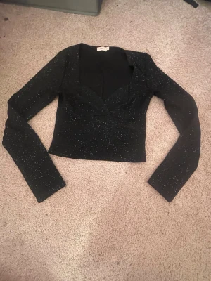 Glittrig svart långärmad topp - Snygg svart topp med lång ärm och glittrigt material. Toppen har en markerad v-ringning och croppad passform som ger en trendig look. Perfekt för dig som vill sticka ut lite extra. Inget synligt märke på plagget.