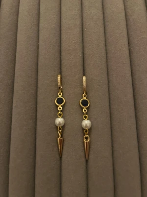 Multiple drop örhängen med’pärlor - Snygga örhängen i guldton med svart detalj, vit pärla och en spetsig konformad hängande del. Örhängena har en modern design med flera olika material och former som ger en cool och trendig look.