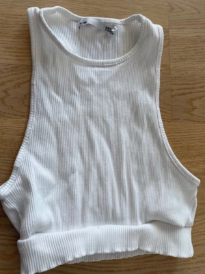 Vit ribbad croppad linnetopp - Snygg vit croppad linnetopp i ribbat material. Toppen är ärmlös med rund halsringning och breda axelband, perfekt för en trendig och avslappnad stil.