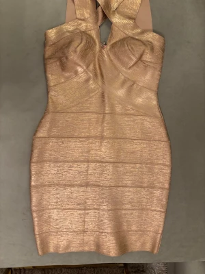 Guldig kort bandageklänning - Säljer en snygg guldig kort bandageklänning med korsade axelband och markerad byst. Klänningen har en kroppsnära passform och ett glansigt material som ger en lyxig känsla. Perfekt för dig som vill sticka ut och glänsa.