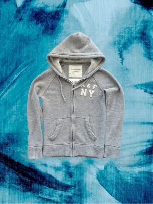 Grå hoodie från Abercrombie & Fitch - Riktigt fet sällsynt grå zip-hoodie från Abercrombie & Fitch med broderad text 'A&F NY' på bröstet. Hoodien har dragsko i huvan, två framfickor och ribbade muddar. Tillverkad i mjukt bomullsmaterial som känns riktigt skönt mot huden.