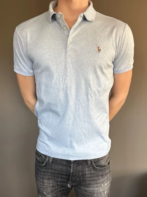 Ljusblå pikétröja från Ralph Lauren - Klassisk ljusblå pikétröja från Ralph Lauren med korta ärmar och krage. Tröjan har knappar framtill och den ikoniska broderade loggan på bröstet. Perfekt för en avslappnad men stilren look. Nypris 2000kr+         Aldrig använd!