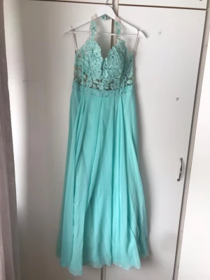 Ljusblå långklänning med spetsdetaljer - Elegant ljusblå långklänning med spetsdetaljer upptill och halterneck. Kjolen är i ett flytande, tunt material som ger ett drömmigt intryck. Överdelen har vackra broderier och transparenta partier, vilket ger en romantisk känsla.