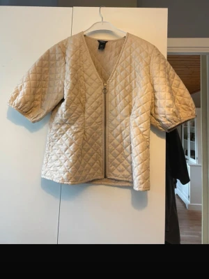 Beige quiltad jacka med puffärm - Quiltad beige jacka med korta puffärmar och dragkedja framtill. Jackan har ett rutigt mönster och v-ringad hals. Perfekt för dig som vill ha en trendig och lätt jacka till vardagslooken.