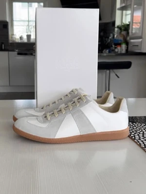 Vita och grå sneakers från Maison Margiela - Snygga sneakers från Maison Margiela i vitt och ljusgrått läder och mocka. Klassisk låg modell med beige snörning och gummisula i brunt. Perfekta för dig som gillar stilren och minimalistisk design. OBS INGEN BOX MEDFÖRLJER
