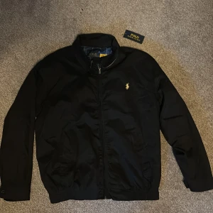 Jacka från Polo Ralph Lauren - Svart jacka från Polo Ralph Lauren med klassisk gul broderad logga på bröstet. Jackan har dragkedja framtill, ståkrage och två sidofickor. Insidan är fodrad med rutigt tyg. Passar S/M i storlek.