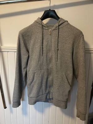 Grå zip hoodie med fickor - En stilren grå hoodie med dragkedja framtill och två stora fickor. Tröjan har huva med dragsko och ribbade muddar vid ärmar och nederkant. Perfekt för en avslappnad look och enkel att matcha med det mesta. Materialet känns mjukt och bekvämt. Pris kan diskuteras vid snabb affär.