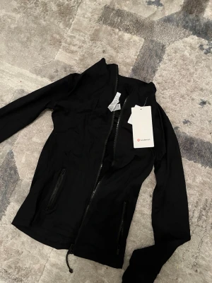 Svart lululemon ziptröja - Snygg svart ziptröja från Lululemon med hög krage och dragkedja framtill. Tröjan har två fickor med dragkedja och är tillverkad i ett stretchigt funktionsmaterial som passar perfekt till träning eller chill. Stilren design med diskret logga.