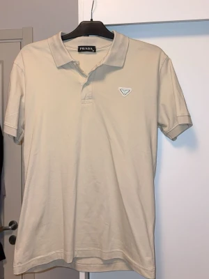 Beige pikétröja från Prada - Stilren beige pikétröja från Prada med klassisk krage och knappslå framtill. Tröjan har korta ärmar och en diskret Prada-logga på bröstet. Perfekt för en clean och avslappnad look.passar både S-M