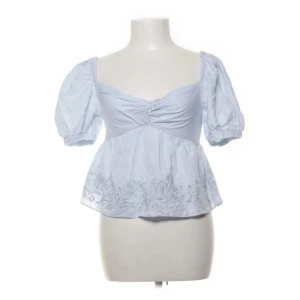 Ljusblå broderad blus med puffärm - Supersöt ljusblå blus med puffärmar och broderade detaljer längst ner. Toppen har en v-ringad urringning med lätt rynkning framtill och en luftig, något croppad passform. Perfekt för dig som gillar romantisk och trendig stil.