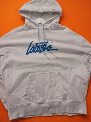 Vit hoodie från Lacoste med blå logga - Snygg vit hoodie från Lacoste med stor blå logga i graffiti-stil på bröstet. Klassisk känguruficka framtill, huva med dragsko och ribbade muddar. Perfekt för en avslappnad streetwear-look.