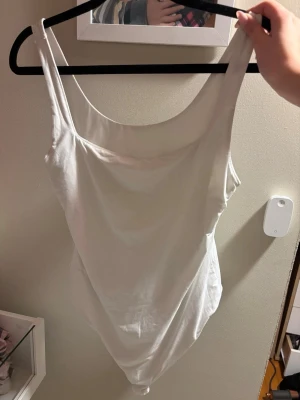 Vit basic body med breda axelband - Säljer en enkel och stilren vit body med breda axelband och rundad urringning. Perfekt att matcha med jeans eller kjol för en clean look. Tillverkad i mjukt stretchigt material som sitter skönt mot kroppen. Passar S och Xs väldigt stretchig!