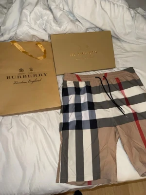 Rutig Burberry shorts med snörning - Säljer ett par klassiska Burberry shorts med ikoniskt rutigt mönster i beige, svart, vit och röd. Shortsen har elastisk midja med svart snörning och är tillverkade i ett mjukt bomullsmaterial. Perfekta för dig som vill ha en stilren och lyxig look.