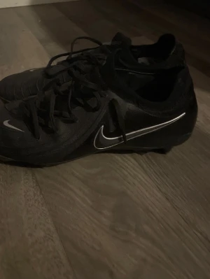 Svarta Nike fotbollsskor med snörning - Säljer ett par svarta fotbollsskor från Nike med klassisk låg profil och vita detaljer på sidorna. Skorna har snörning och är tillverkade i syntetmaterial för bra grepp och kontroll på planen. Perfekta för dig som vill ha stilrena och funktionella skor till matchen.