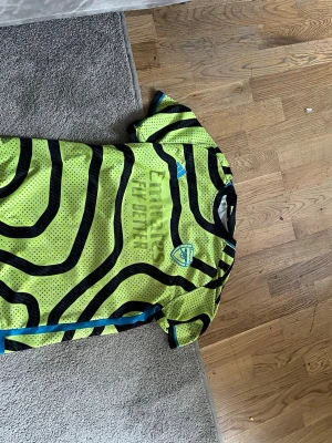 Arsenal neon matchtröja Adidas - Säljer en Arsenal matchtröja från Adidas i neon-gult med svarta och blå detaljer. Tröjan har korta ärmar, sopiga svarta mönidringar och klubbmärke sop Arsenal HM på bröstet. “Emirates Fly Better” tryckt på framsidan. Perfekt för fotbollsträning.