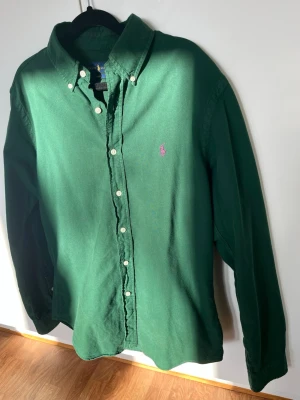 Grön skjorta från Ralph Lauren - Klassisk grön skjorta från Ralph Lauren i slim fit-modell. Skjortan har långa ärmar, knappar framtill och en lila broderad logga på bröstet. Tillverkad i mjuk bomull och perfekt för en clean och stilren look. Storlek M.