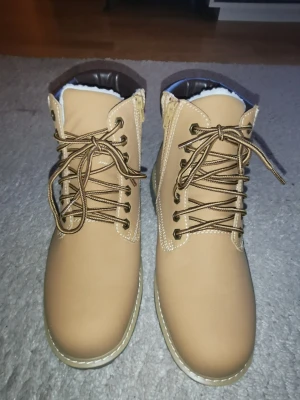 Beige kängor med snörning och dragkedja - Snygga beige kängor med bruna detaljer, snörning framtill och dragkedja på sidan. Grov sula för bra grepp och vadderad insida. Tillverkade i syntetmaterial med kontrastsömmar. Perfekta för höst och vinter.