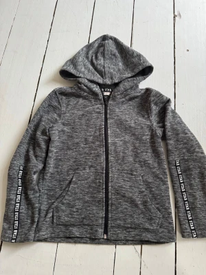 Gråmelerad hoodie fleece - Snygg gråmelerad hoodie från HM med dragkedja framtill och huva. Ärmarna har textdetaljer längs sidorna och tröjan har två fickor framtill. Tillverkad i mjukt och bekvämt material som passar perfekt till en avslappnad stil.