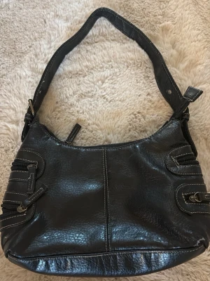 Svart skinnväska - Svart skinnväska. Förmodligen fejkskinn, fått av mormor. Två små fickor på vardera sida av väskan och en liten ficka på insidan av väskan. Dragkedja. 