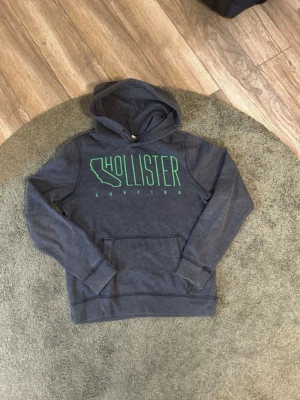 Mörkgrå Hollister hoodie  - Mörkgrå hoodie från Hollister med grön text och Kalifornien-tryck på bröstet. Klassisk känguruficka framtill och huva. Skön och avslappnad passform, perfekt till chill dagar. Tillverkad i mjukt bomullsmaterial.