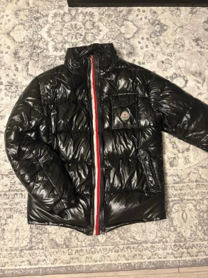 Svart glansig dunjacka från Moncler - Säljer en svart glansig dunjacka från Moncler med hög krage och rödvit rand längs dragkedjan. Jackan har en bröstficka med lock och Moncler-logga, samt två sidofickor. Perfekt för kalla vinterdagar och riktigt snygg streetstil.