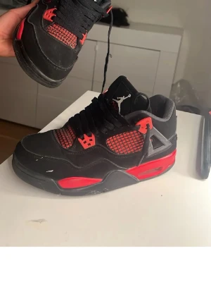  Air Jordan 4 sneakers - Säljer ett par Air Jordan 4 sneakers i svart och rött med meshdetaljer på sidorna och klassisk Jumpman-logga på plösen. Skorna har svarta skosnören, röda accenter och en robust sula med synlig luftdämpning. Materialet är en mix av mocka och textil.