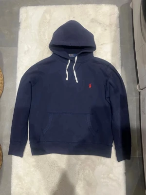 RL hoodie - Säljer en mörkblå hoodie från Polo Ralph Lauren med klassisk huva och vita snören. Tröjan har en röd broderad logga på bröstet och en stor magficka. Materialet är mjukt och bekvämt, perfekt för chill dagar. Stilen är enkel och tidlös. ( använd 2 gånger)