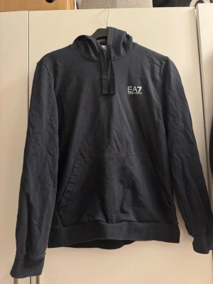 EA7 Emporio Armani hoodie  - EA7 Emporio Armani hoodie i färgen dark blue med silverlogga, storlek M.  Använd max 2 gånger och är som nyskick. Inga hål, inga fläckar och inga synliga tecken på användning.  Nypris ca 500 kr. Säljs då den inte kommer till användning.                   Katt finns i hemmet.