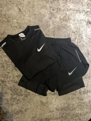 Svart träningsset från Nike - Säljer ett svart träningsset från Nike med t-shirt och shorts. Båda plaggen har vita Nike-loggor och reflekterande detaljer. Materialet är lätt och andas, perfekt för löpning eller gym. Strl S, passar dig runt 160 lång, hör gärna av dig vid funderingar kring mått eller annat!