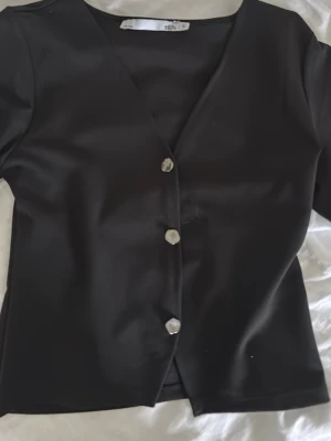 Svart kortärmad topp med knappar - Svart topp med v-ringning och korta ärmar från 15A. Toppen har dekorativa silvriga knappar framtill och är gjord i ett slätt syntetmaterial. Passformen är normal och den är enkel att matcha med olika outfits.