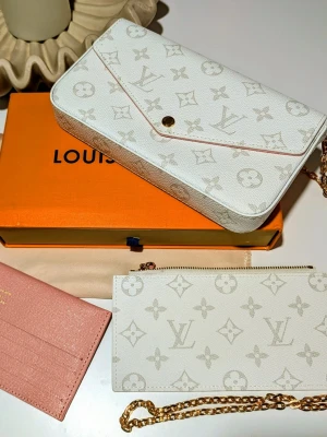 Iouis Vuitton handväska  - Väskan: En rektangulär kuvertväska i vitt med ett ljusgrått monogram-mönster. Den har ett spetsigt lock (envelope-stil) som stängs med en guldfärgad tryckknapp i mitten. Längs kanten syns en diskret rosa kontrasterande färg. Axelrem: En avtagbar, tunn kedja i guldfärgad metall som är fäst vid väskans sidor.