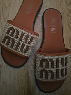 Bruna sandaler  - Snygga sandaler med flätad beige rem och brunt broderat logotypmönster. Sulan är brun med svarta kanter och öppen tå. Perfekta för en trendig och avslappnad look.