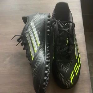 Adidas F50 svart/gul fotbollssko - Säljer ett par Adidas F50 fotbollsskor i svart med neon-gula detaljer och vita ränder. Skorna har snörning, låg profil och slitstark yttersula med dobbar för bra grepp på konstgräs. Snygg, sportig design med F50-logga på ovansidan. Perfekta för dig som vill sticka ut på planen.
