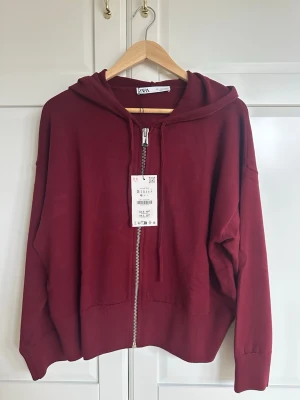 Hoodie med dragkedja från Zara - Snygg vinröd finstickad hoodie från Zara med dragkedja framtill och luva. Modellen är oversized med nedhasade axlar och ribbade muddar. Tillverkad i mjukt material som känns skönt mot huden. Perfekt för en avslappnad och trendig look. Lite dåligt ljus på flera av bilderna, men skulle säga att färgen på näst sista bilens är mest lik hur den ser ut i verkligheten. Aldrig använd och prislappen sitter kvar. Nypris: 470kr