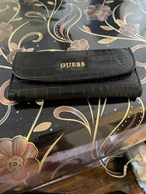 Svart plånbok i croco-mönster Guess - Snygg svart plånbok från Guess med croco-mönstrad yta och guldfärgad logga på framsidan. Plånboken har en avlång form och stängs med lock. Perfekt för dig som vill ha en stilren accessoar med lyxig känsla.