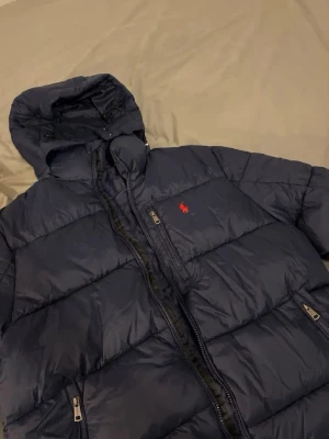 Ralph Laurent  - Snygg mörkblå pufferjacka från Polo Ralph Lauren med röd broderad logga på bröstet. Jackan har huva, två dragkedjeförsedda fickor och quiltad design. Perfekt för kalla dagar och riktigt skön att bära. Materialet är glansigt och ger en modern look.