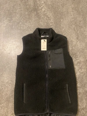 Svart fleeceväst med dragkedja - Mysig svart fleeceväst med hög krage och hel dragkedja framtill. Västen har två fickor i sidorna och en bröstficka med dragkedja i kontrasterande tyg. Perfekt för lager-på-lager och enkel att matcha till olika outfits.