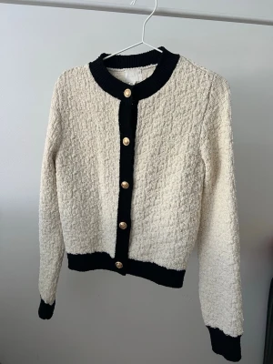 Krämvit kofta med svarta detaljer - Stilren krämvit kofta med svarta ribbade kantermanchetter och guldknappar framtill. Koftan har en ~croppad~ passform, rund halsringning och lång ärm. Materialet är strukt reclaimat och ger en lyxig känsla. Perfekt för dig som vill ha en chic och trendig look.