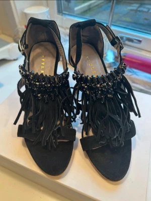 Svarta pumps med fransar från Stylesnob - Säljer ett par versatile svarta pumps från Stylesnob i modellen France. Skorna har öppen tå, hög klack i trälook och snygga fransdetaljer med svarta pärlor framtill. Rem runt ankeln med spänne och ovandel i mockaimitation. Perfekta för dig som vill sticka ut!