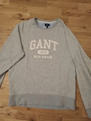Grå sweatshirt från GANT XL - Klassisk grå sweatshirt från GANT med tryck 'GANT 1949 NEW HAVEN' på bröstet. Tröjan har rund halsringning, långa ärmar och ribbade muddar. Perfekt för en avslappnad och stilren look.