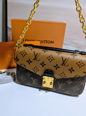 Iouis Vuitton handväska  - Butikspriset är 26 000kr.Detta är en rektangulär axelremsväska med en klassisk och strukturerad silhuett. Väskan har ett lock som smalnar av till en V-formad flik framtill, vilket ger den ett elegant och symmetriskt uttryck. Material: Väskan är tillverkad i ett mönstrat material med en tydlig textur. 