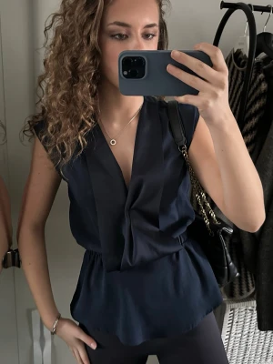 Mörkblå omlottopp med v-ringning - Snygg mörkblå omlottopp med v-ringning och resår i midjan som ger en fin form. Toppen är ärmlös och har en lätt peplum-effekt nedtill. Perfekt att styla med jeans eller kjol för en trendig look.