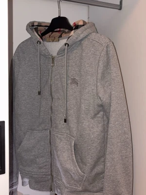 Grå zip-hoodie från Burberry - Snygg grå hoodie med dragkedja från Burberry. Klassisk huva med dragsko och ikoniskt rutigt foder på insidan. Mjuk bomullsmix, två fickor framtill och diskret logga broderad på bröstet. Perfekt för en chill och stilren look.