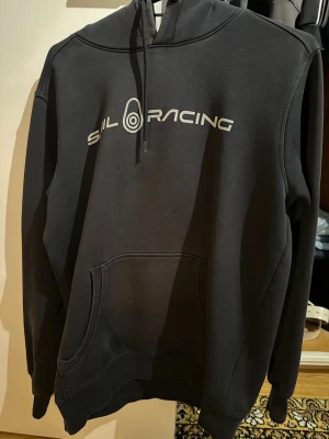 Svart hoodie SIL RACING - Svart hoodie från SIL RACING med vit logga och text på bröstet samt en mindre logga på huvan. Klassisk känguruficka framtill och justerbar dragsko i huvan. Mjuk bomullsblandning och ribbade muddar vid ärmslut och nederkant.