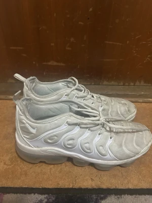 Nike vita Air Max Plus sneakers - Nike Air Max Plus sneakers i helvitt med ikoniska vågformade detaljer och synlig Air-sula. Ovandelen är i textil och syntet, med vita snören och diskret Swoosh-logga på sidan. Skorna har en sportig och futuristisk look som sticker ut.