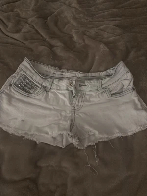 Vita jeansshorts med nitar - Säljer ett par vita jeansshorts med råa kanter och coola nitar på ena fickan. Shortsen har låg midja, klassiska fickor och bälteshällor. Perfekta för varma dagar och en avslappnad stil.