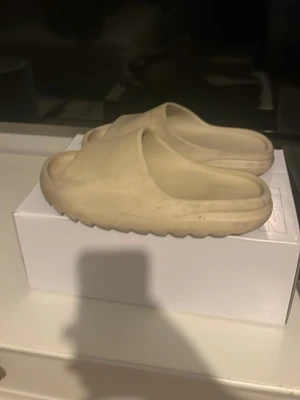 Yezzy slides Beige  - Säljer ett par beige slip-on tofflor med chunky sula och öppen tå. Tofflorna är tillverkade i ett mjukt och flexibelt syntetmaterial som ger en skön känsla. Perfekt för chill dagar hemma eller när du vill ha något bekvämt på fötterna.