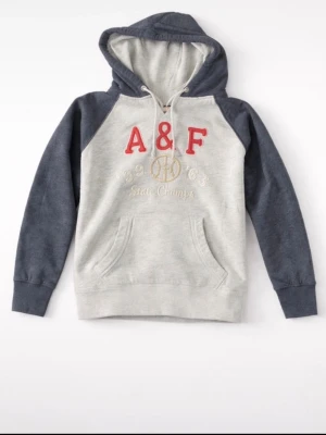Grå och mörkblå hoodie från A&F - Snygg hoodie från Abercrombie & Fitch i grått med mörkblå ärmar och huva. Stor röd A&F-logga framtill och broderad text i guld. Klassisk känguruficka och dragsko i huvan. Perfekt för en avslappnad streetstyle.