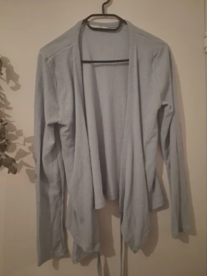 Ljusblå ribbad kofta - Söt ljusblå kofta i ribbat material med långa ärmar och knytband framtill. Koftan har en öppen design utan knappar och faller snyggt längs kroppen. Perfekt att slänga över en topp för en avslappnad look.