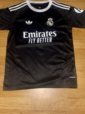 Svart Real Madrid Adidas tröja - Snygg svart Real Madrid tröja från Adidas med vita detaljer och tryck. Tröjan har klubbmärke, Adidas-logga och Emirates Fly Better-tryck på bröstet. Tillverkad i lätt och funktionellt material med Aeroready-teknologi. Perfekt för fotbollsträning eller streetwear.
